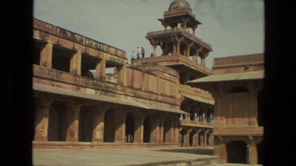 1974 India travel frame showing a palace courtyard with colonnaded walls, a raised pavilion, roofline shadows, and distant visitors on the structure.