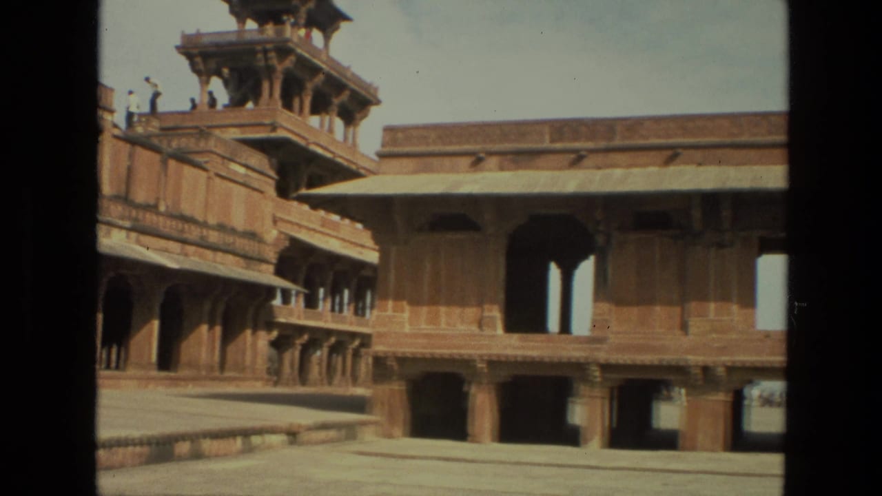 1974 India travel footage frame showing a Mughal-era palace courtyard with red sandstone arches, upper walkway visitors, and a towered pavilion.