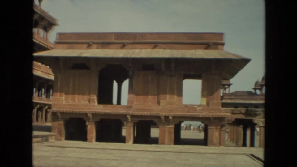 1974 8mm frame from India showing a broad sandstone palace facade with open arches, deep shadows, and courtyard paving.
