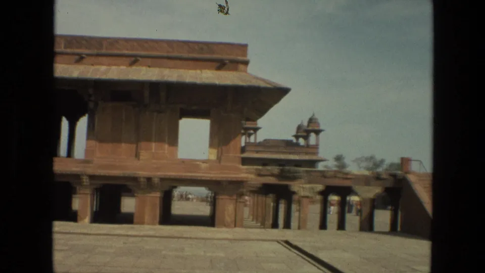 1974 India home-movie frame showing a sandstone palace courtyard, open archways, roofline details, and distant visitors near the far passage.