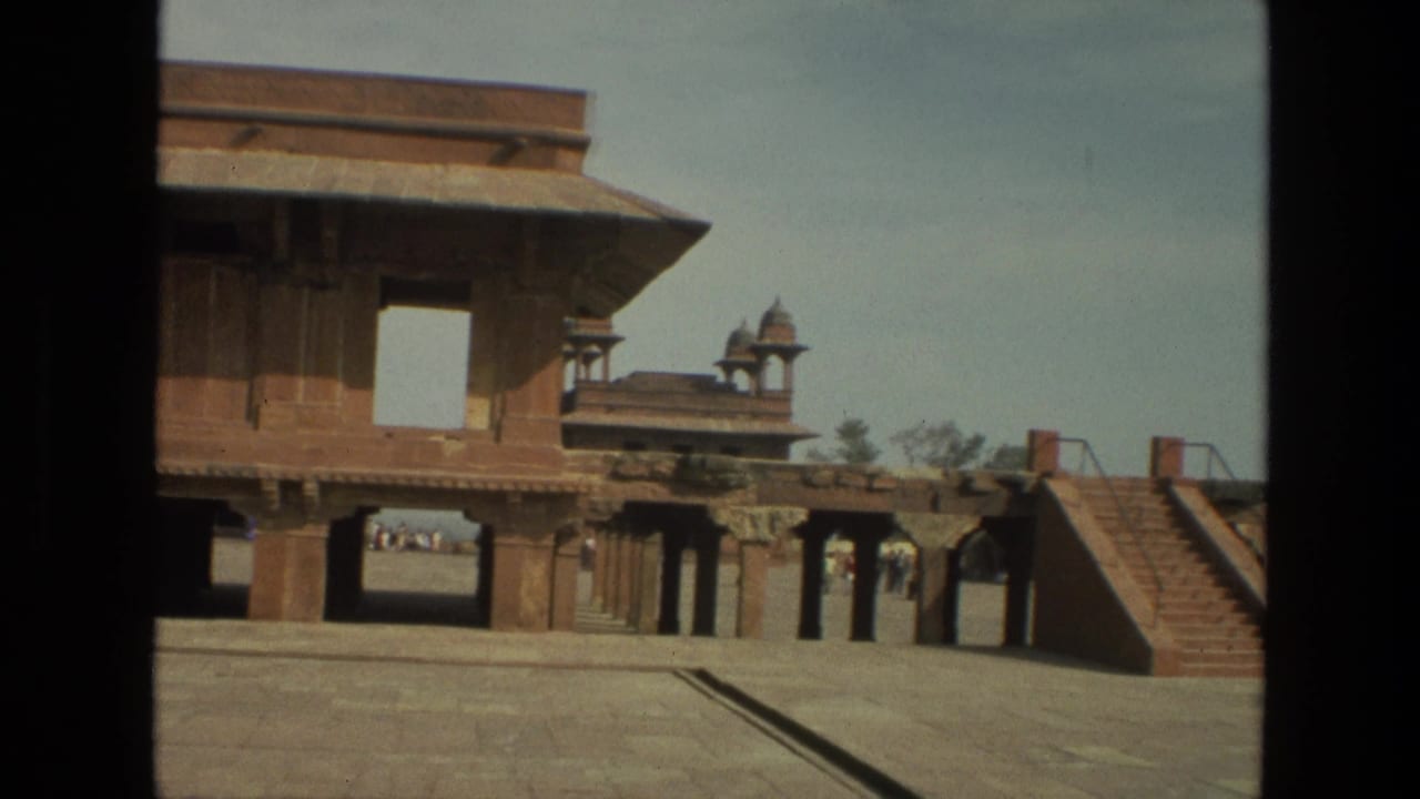 1974 India home-movie frame showing a courtyard, carved stone architecture, steps, and small distant visitors.