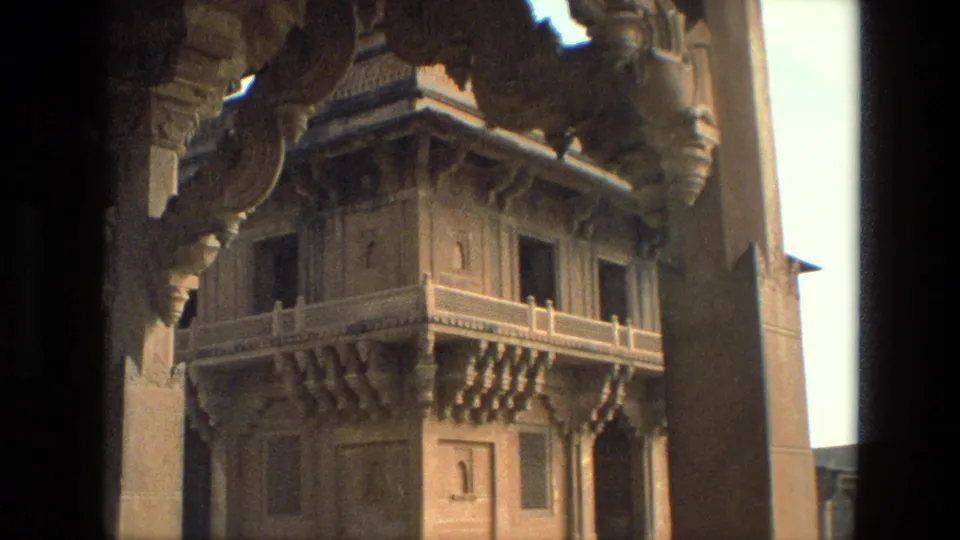 1974 India home-movie frame with dark pillar shadows framing an ornate temple balcony, carved brackets, windows, and sunlit stone surfaces.