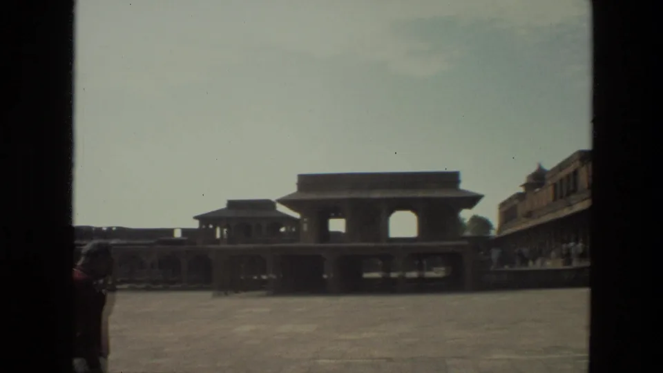 1974 India travel frame showing a broad palace courtyard, arched structures, and a lone visitor partly visible near the left edge.