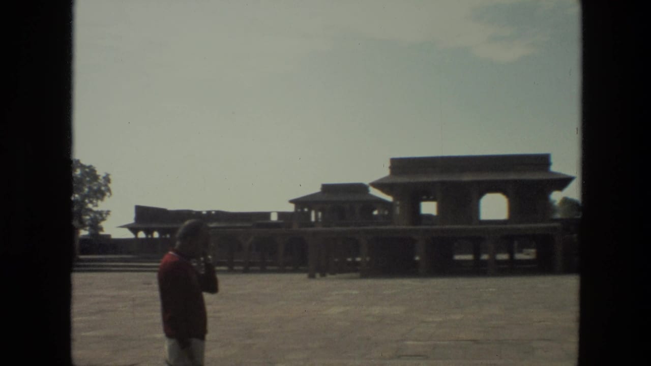 1974 India home-movie frame showing a man in a red shirt near a large paved courtyard and arched historic architecture.