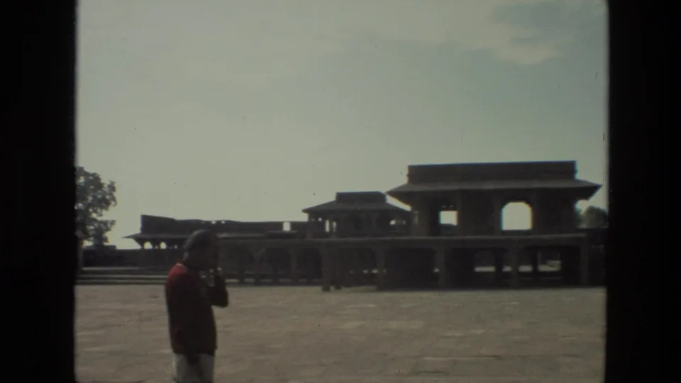 1974 India home-movie frame showing a man in a red shirt near a large paved courtyard and arched historic architecture.