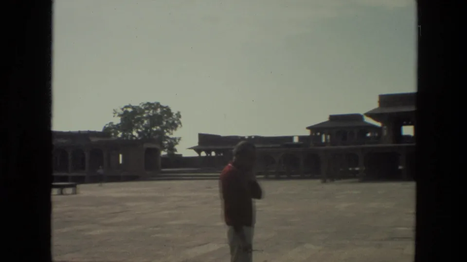 1974 India 8mm frame with a man standing in profile before open stone architecture and a mostly empty courtyard.