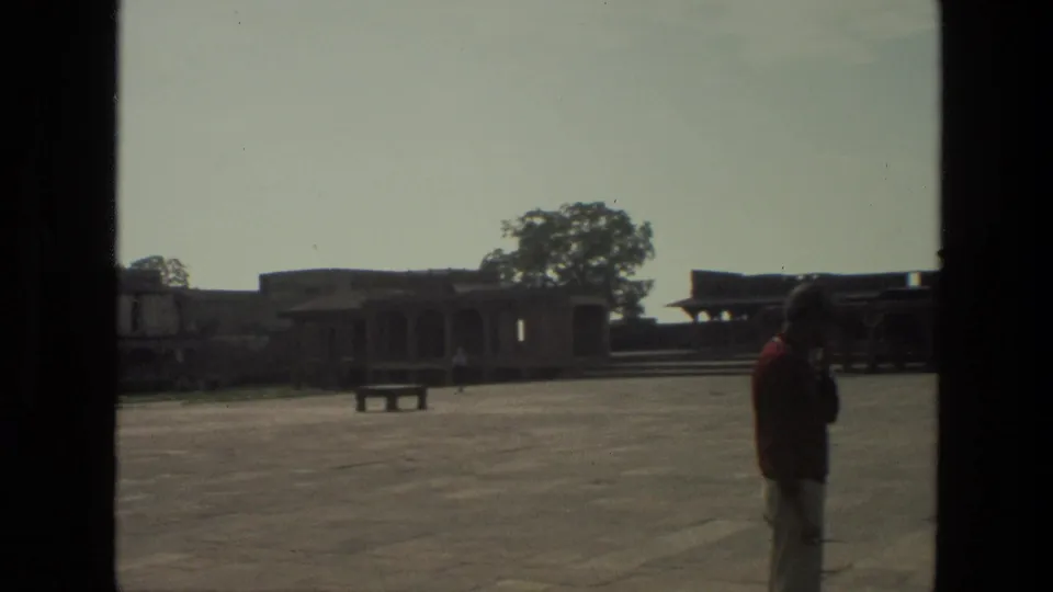 1974 India travel-film frame showing a red-shirted man in a large courtyard with a bench, trees, and historic stone structures.