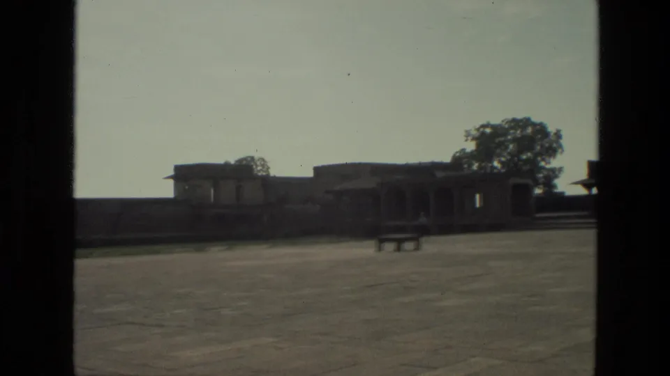 1974 India 8mm frame after the man exits, leaving an empty courtyard, bench, trees, and historic architecture in view.
