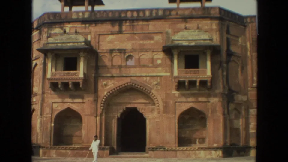 1974 India frame with a person in white crossing the courtyard before a red sandstone building with arched entrances and balcony windows.