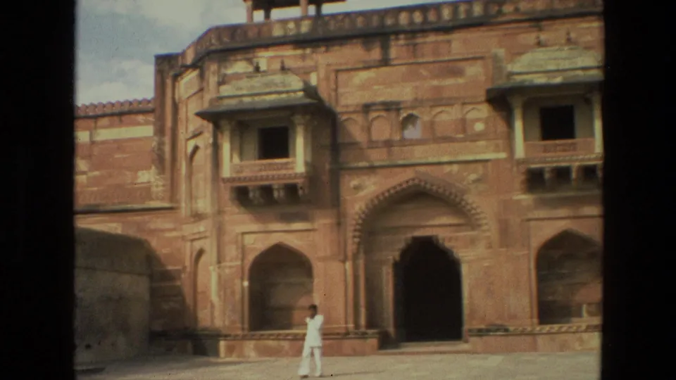 1974 India 8mm frame showing a person in white near a dark arched entrance of a historic red sandstone building.