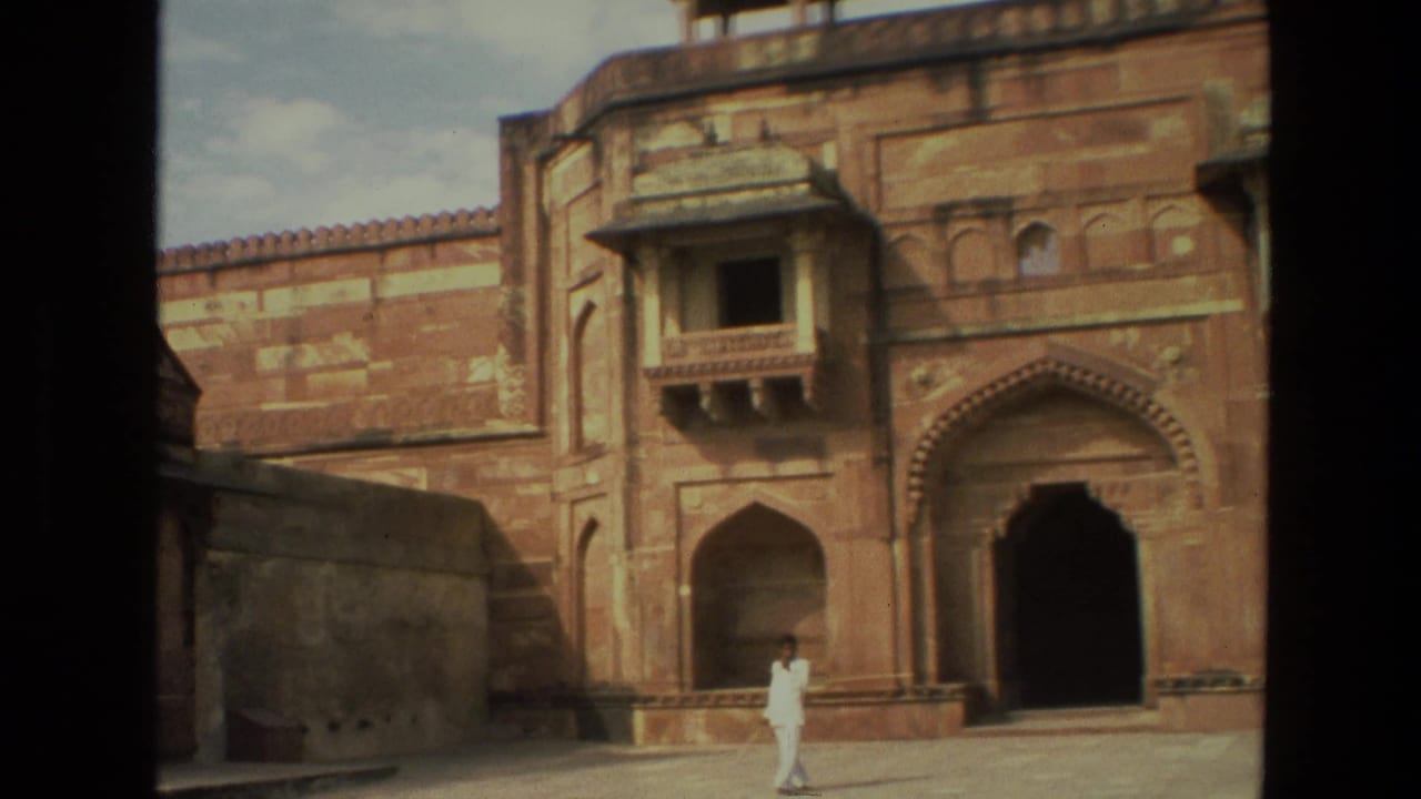 1974 India 8mm frame showing a man in white clothing crossing palace grounds in front of red sandstone arches and a projecting balcony.