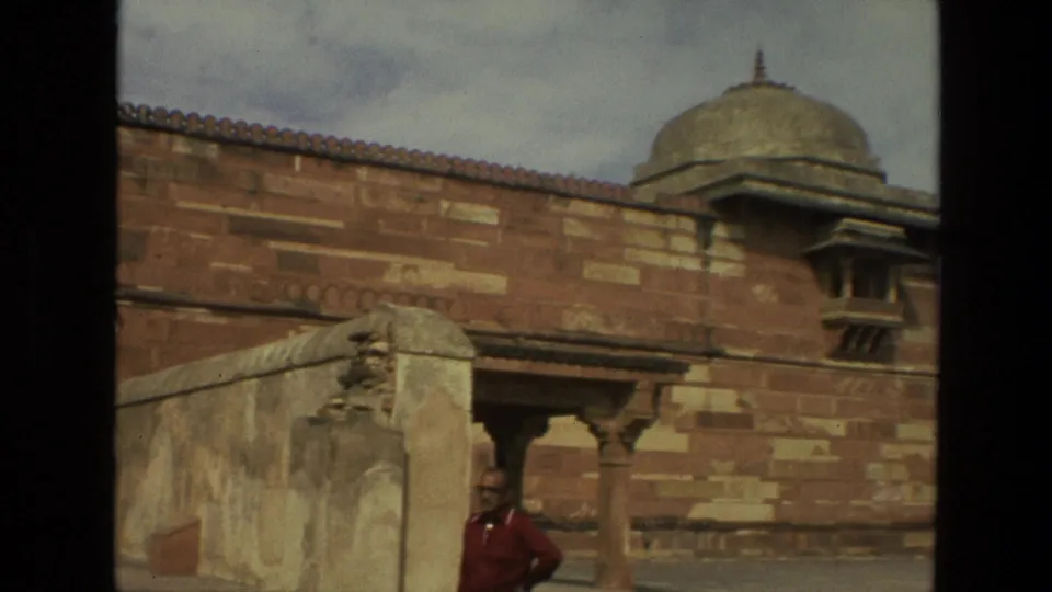 1974 India 8mm view of a red sandstone wall, domed pavilion, arched openings, and an adult visitor standing near the courtyard edge.