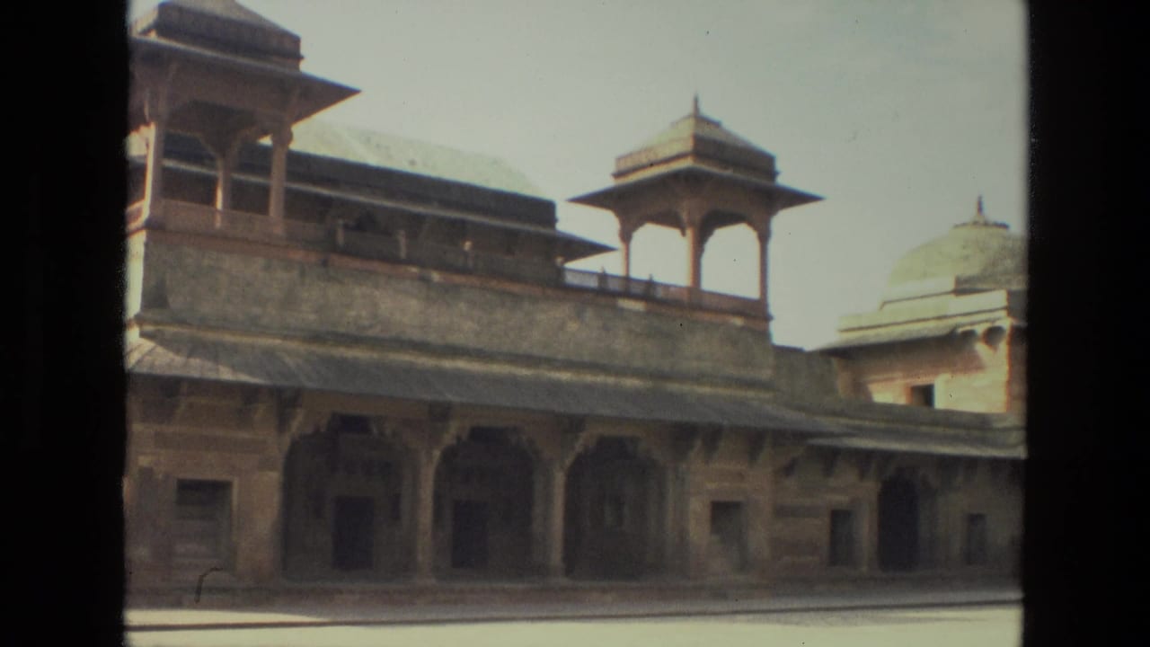 1974 India 8mm frame of a historic sandstone monument with domed pavilion, arched colonnade, shaded openings, and bright courtyard light.