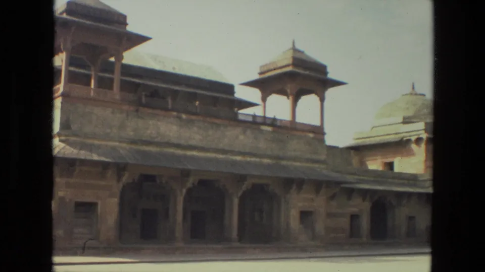 1974 India 8mm frame of a historic sandstone monument with domed pavilion, arched colonnade, shaded openings, and bright courtyard light.