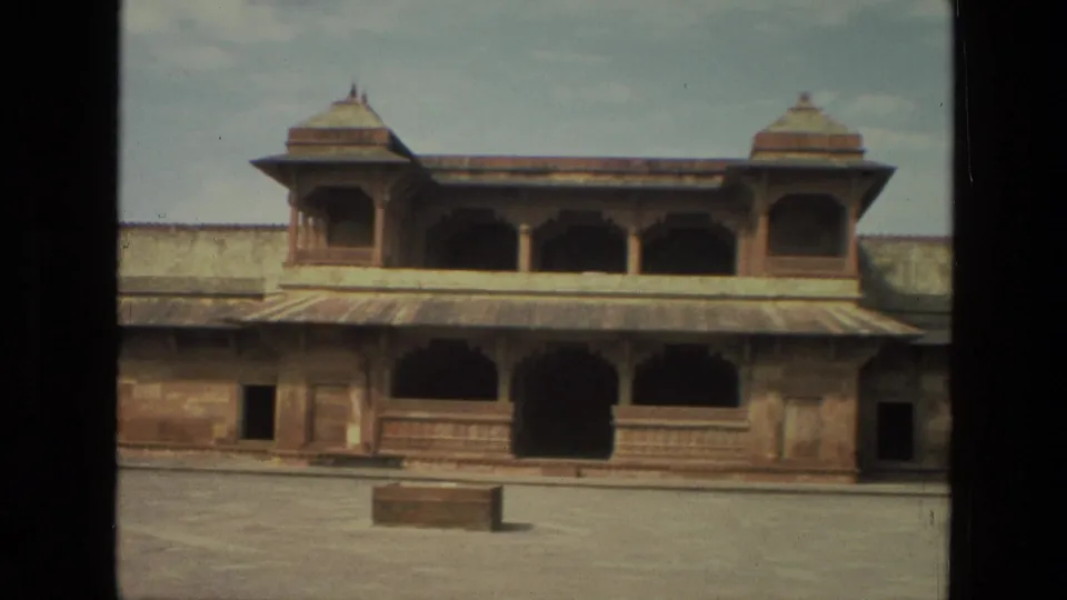 1974 India 8mm frame showing an empty stone courtyard below red sandstone arches, carved railings, and small domed pavilions.