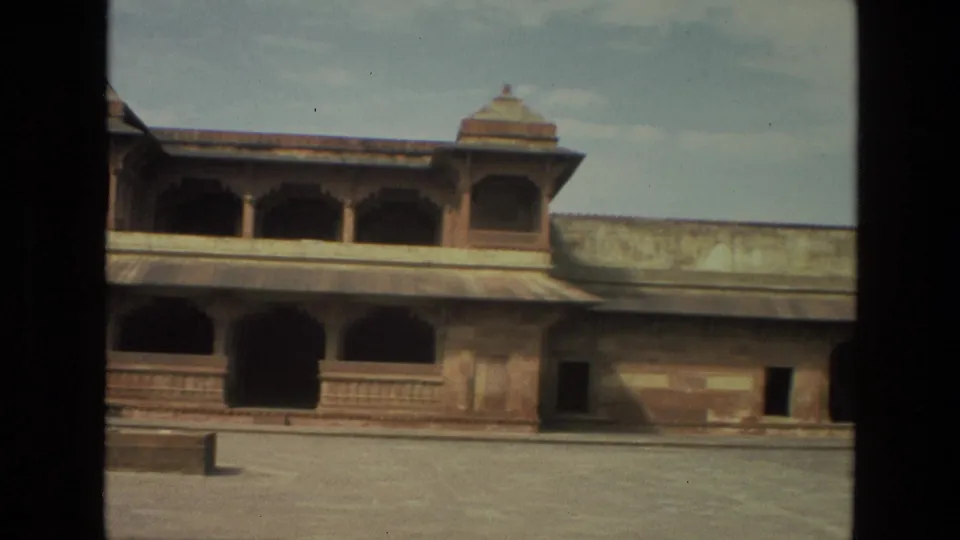 1974 India 8mm frame of a quiet Mughal-era courtyard, with a sandstone arcade, domed roof kiosk, weathered wall, and small rectangular block on paving.