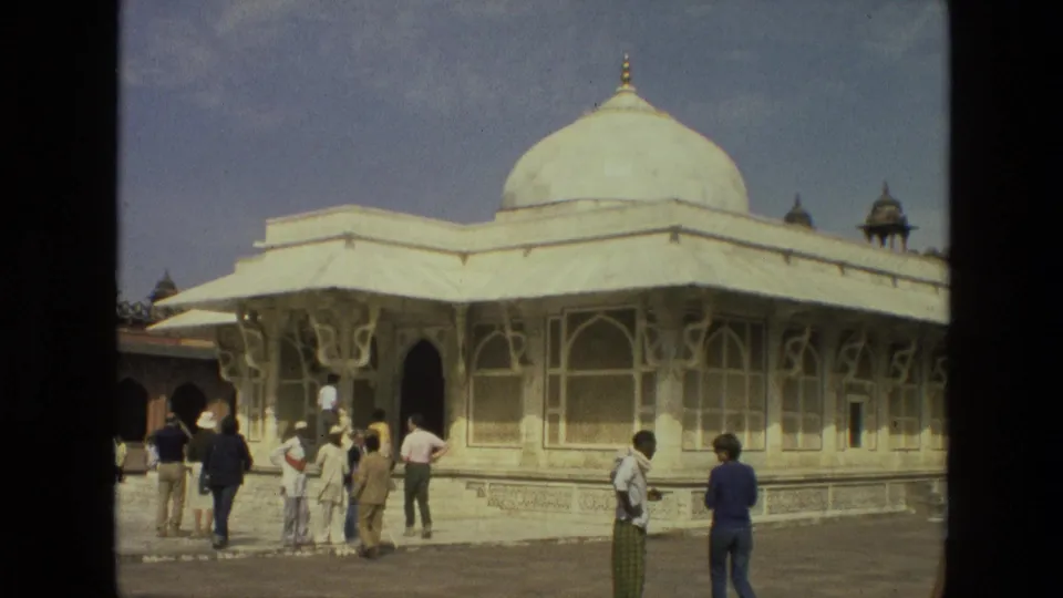 Visitors in 1970s India stand and walk beside a white domed mosque with arched windows and decorative exterior details.