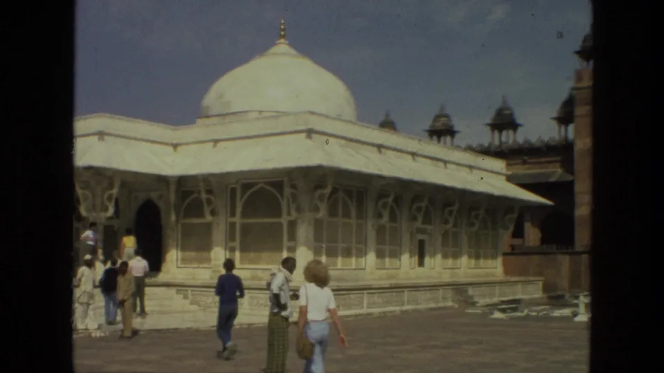 Tourists in a courtyard pass in front of a white domed mosque in India, filmed with warm, grainy 8mm texture.