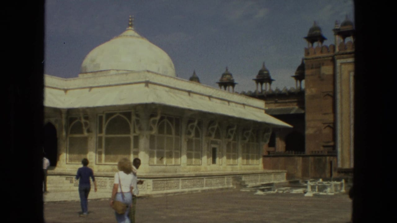 Visitors walk across a sunlit courtyard beside a white domed pavilion and red sandstone gateway.