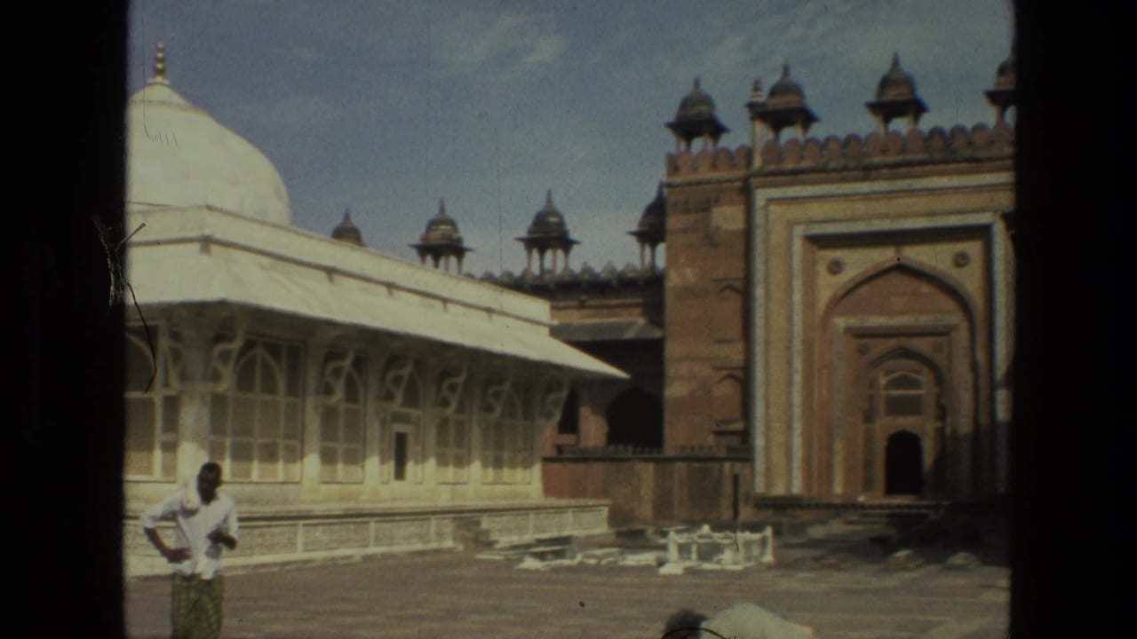 1974 India home-movie frame with a visitor in front of pale arched architecture and a red sandstone gateway with roof pavilions.