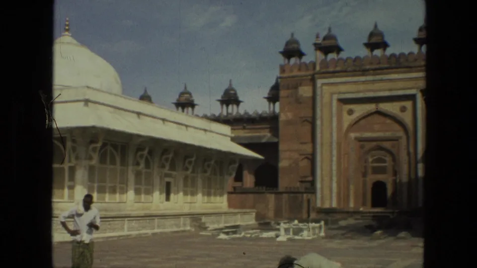 1974 India home-movie frame with a visitor in front of pale arched architecture and a red sandstone gateway with roof pavilions.