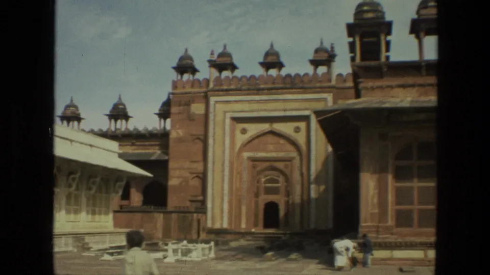Handheld 8mm view of an Indian historic monument courtyard with a red sandstone gateway, pale arched building, and visitors near the foreground.