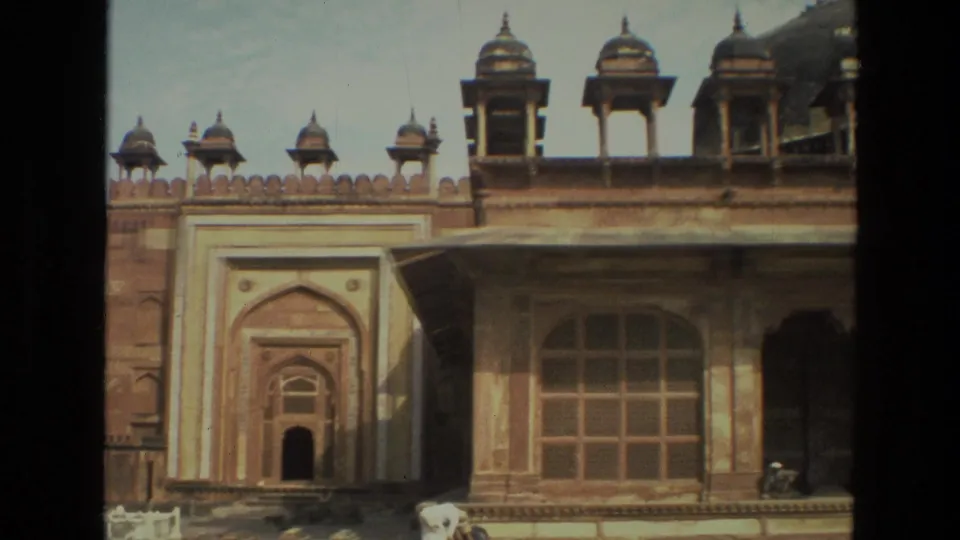 1974 8mm home-movie frame in India showing a historic red sandstone gateway, adjacent pale arched structure, and visitors along the courtyard edge.
