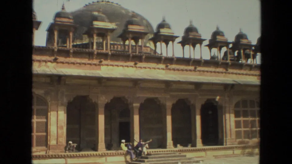 1974 India home-movie view angled across the historic facade, with domes above shaded arches and visitors sitting on steps in the courtyard.