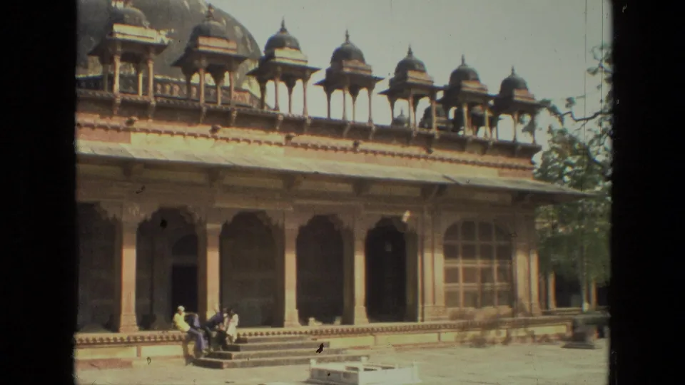 1974 India frame showing the complex across a courtyard, with ornate rooftop kiosks, a shaded arched facade, trees at the edge, and visitors on the steps.