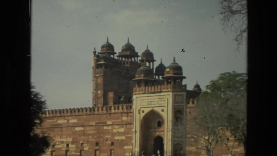 1974 India 8mm frame with a wider view of the fort wall, arched gateway, upper domes, trees, and a small person at the entrance.