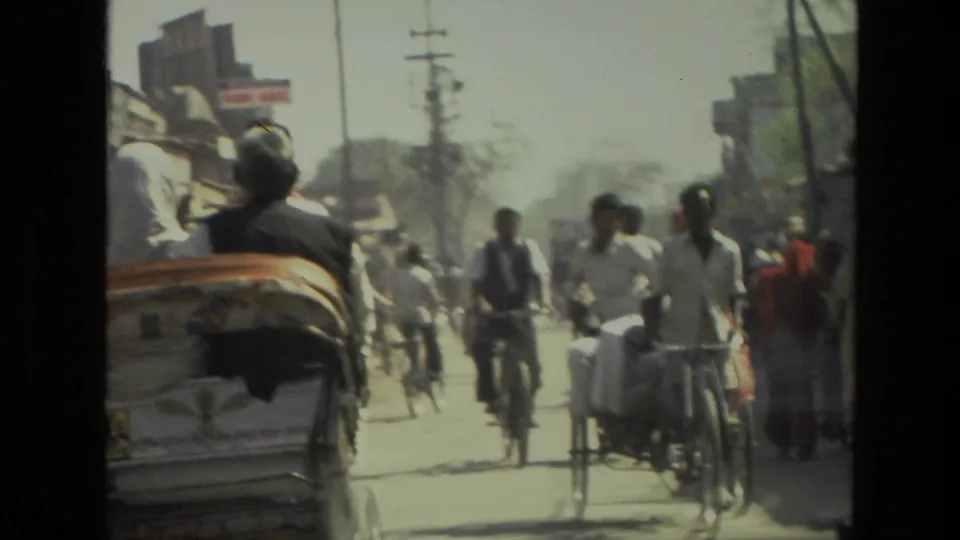 1974 India home-movie frame showing cyclists, pedestrians, buildings, utility poles, and a cart on a dusty town street.