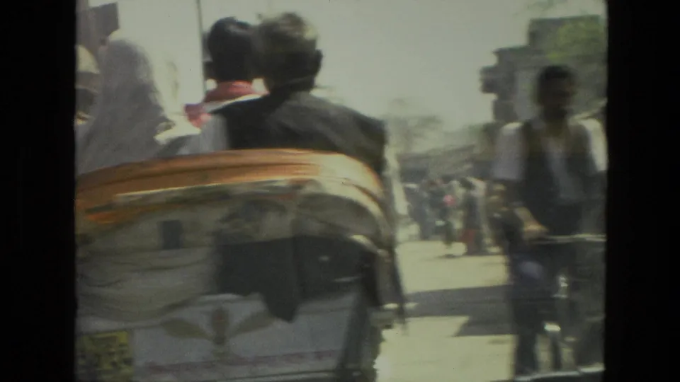 1974 India street scene with the rear of a cart, nearby pedestrians, cyclists, and hazy roadside buildings in bright daylight.