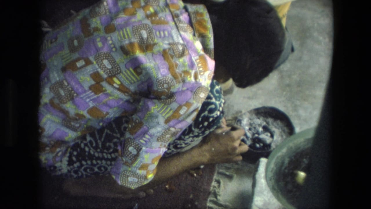 A woman in patterned clothing leans forward over a dark rounded work object beside a basin in the 1974 India indoor craft scene.