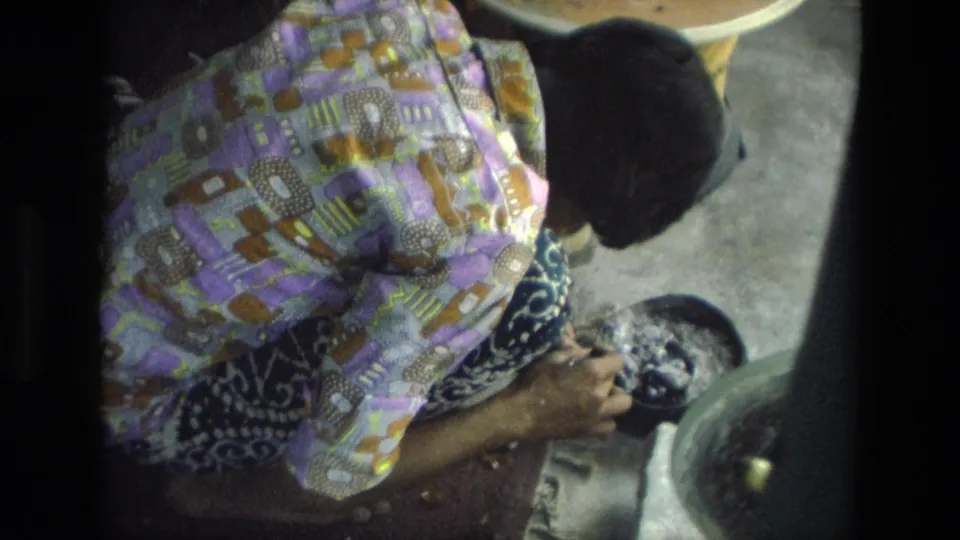 The overhead 8mm frame shows a woman in patterned clothing working by hand near a basin and low table in an indoor setting in India.