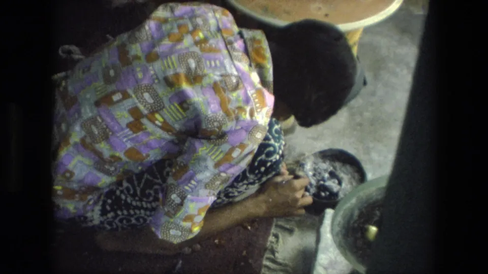 A later frame shows the woman still bent over the same handwork area, with a basin and low round table visible in the 1974 India home-movie scene.