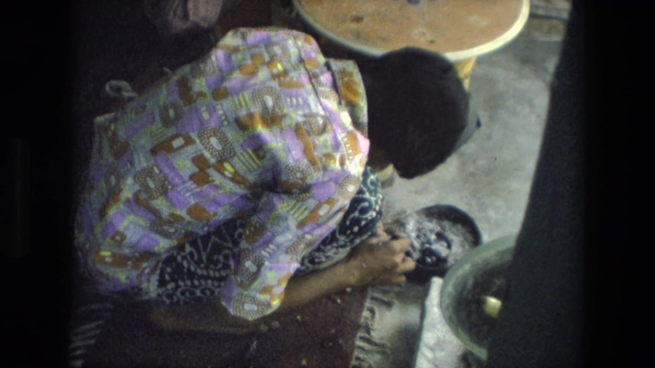 Woman bends over a low work surface while handling a small tool inside a dim room.
