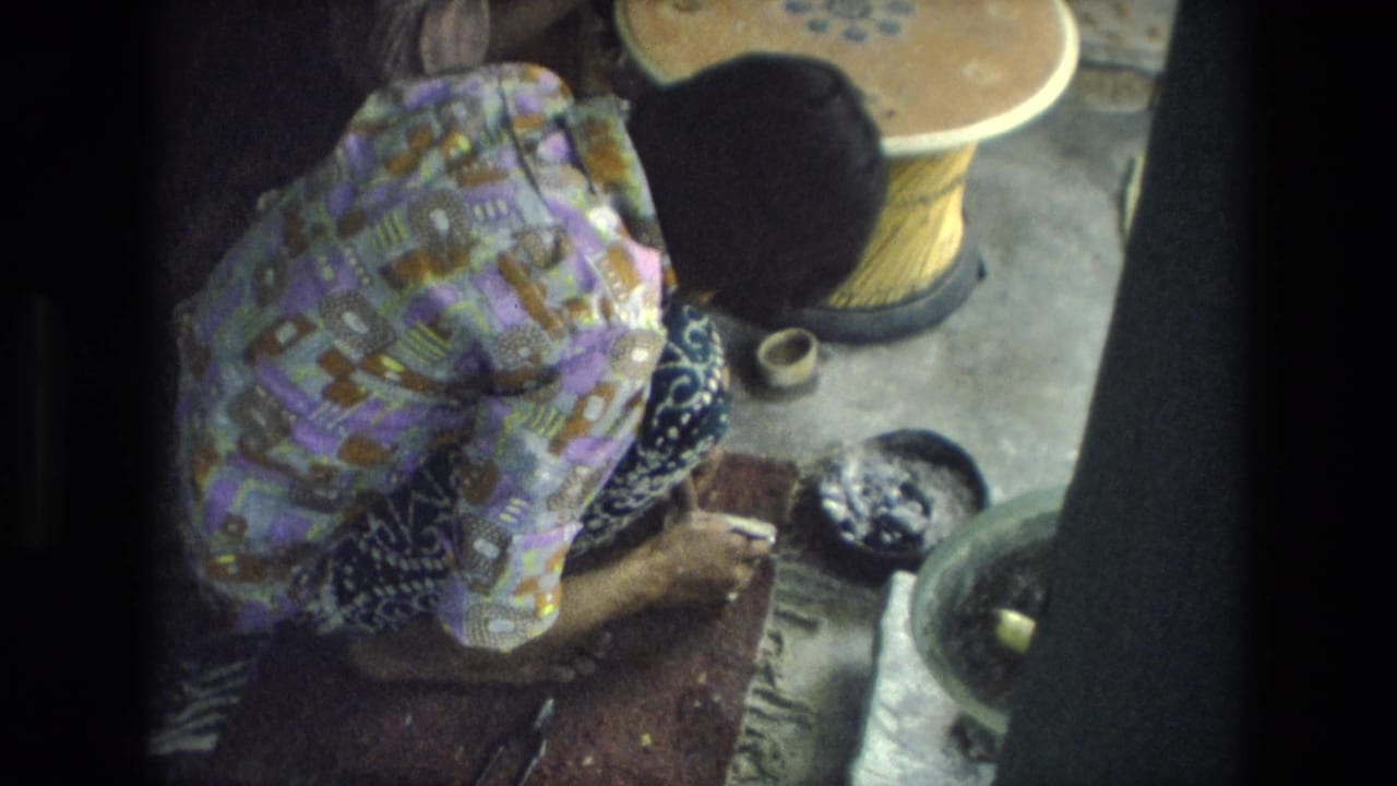 The woman remains focused on handwork beside a bowl, tool, and wooden wheel-like object in the 1974 India home-movie frame.