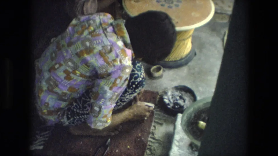 The woman remains focused on handwork beside a bowl, tool, and wooden wheel-like object in the 1974 India home-movie frame.