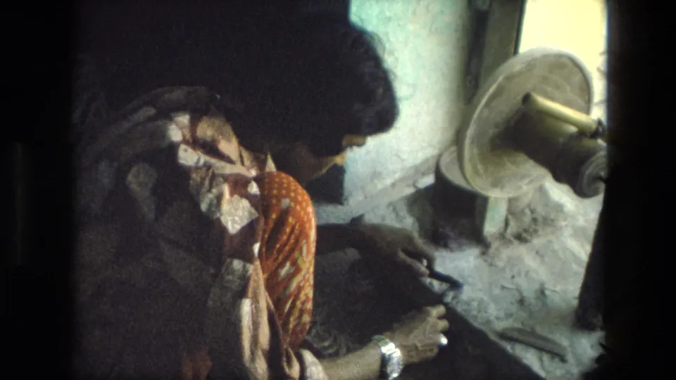 A 1974 India home-movie frame shows the woman seated in shadow beside a spinning wheel-like object, with her hands near her lap.
