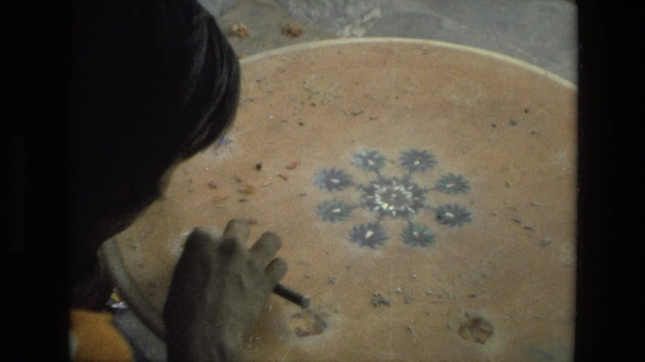 A 1974 India home-movie frame shows a woman working by hand on a round wooden tabletop with a blue floral center.