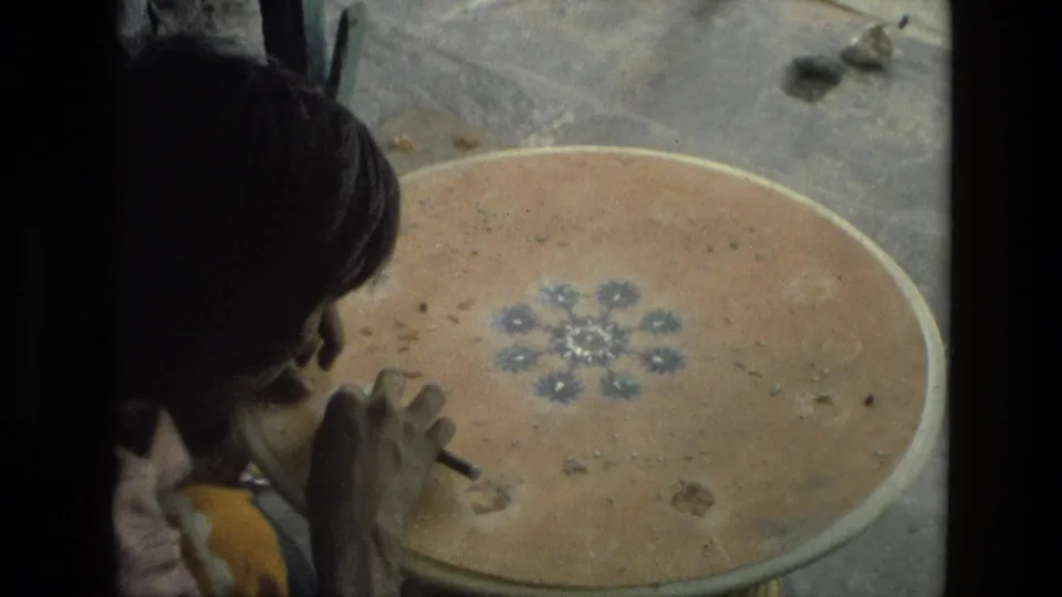 The woman continues detailed handwork on the round wooden table, with the floral design centered in the 1974 India frame.