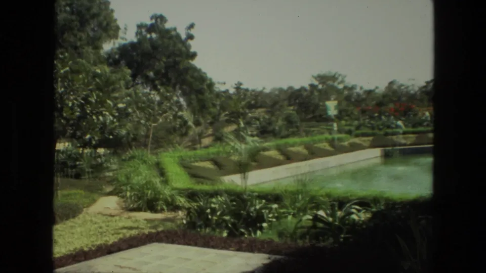 A 1974 India home-movie frame shows a manicured garden pool, low hedges, tropical planting, trees, and pale paving in bright daylight.