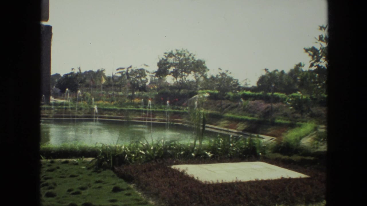Manicured garden with pond fountains, hedges, flowerbeds, and trees in sunlight.