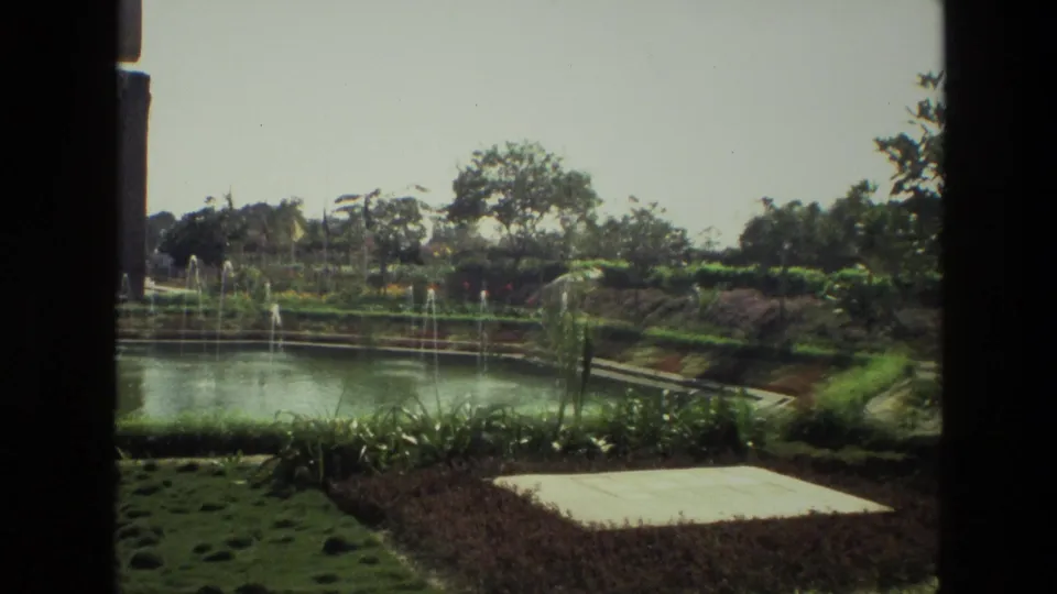 1974 India 8mm frame showing a landscaped garden with pond fountains, clipped hedges, flowerbeds, and trees under bright daylight.
