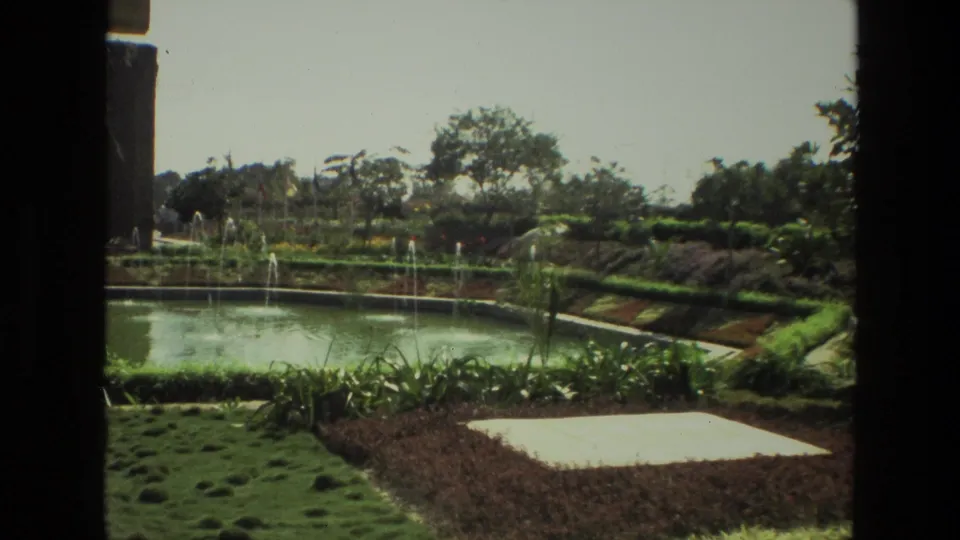 1974 India 8mm frame of a formal garden pond with spraying fountains, trimmed borders, palms, and a pale rectangular paved area.