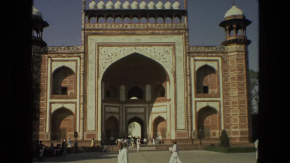 1974 home-movie frame showing the gateway arch, patterned stonework, and tourists in light clothing crossing the foreground.