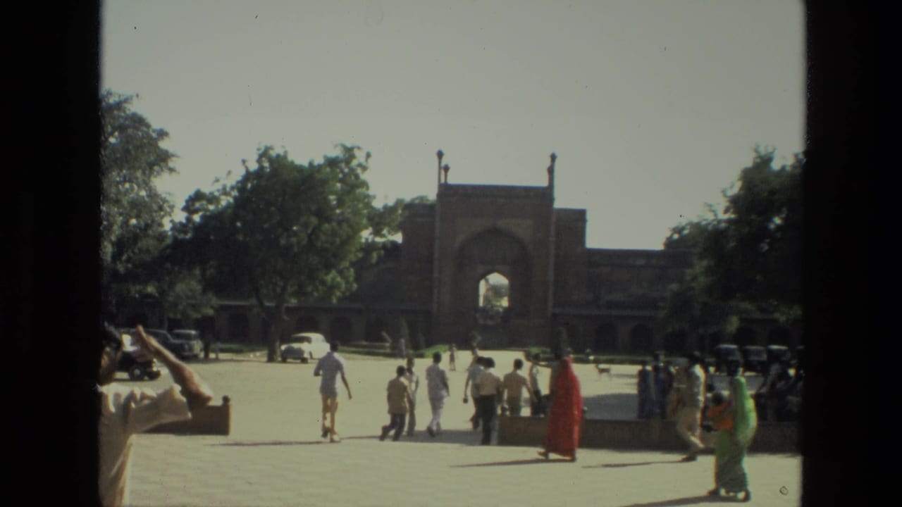 Visitors walk across a sunny courtyard toward a large arched gateway.