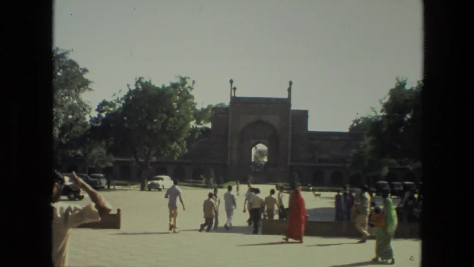 1974 India travel frame showing tourists crossing a courtyard toward a Mughal-style gateway near the Taj Mahal complex.