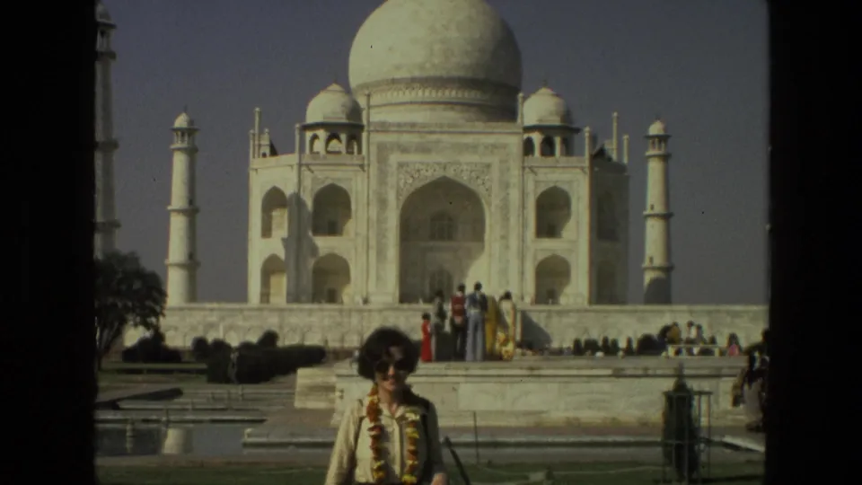 1974 home-movie frame of a woman wearing a floral garland posing before the Taj Mahal, with other visitors on the platform.