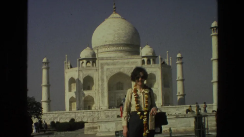 1974 India home-movie frame showing a garlanded woman standing before the Taj Mahal with tourists in the background.